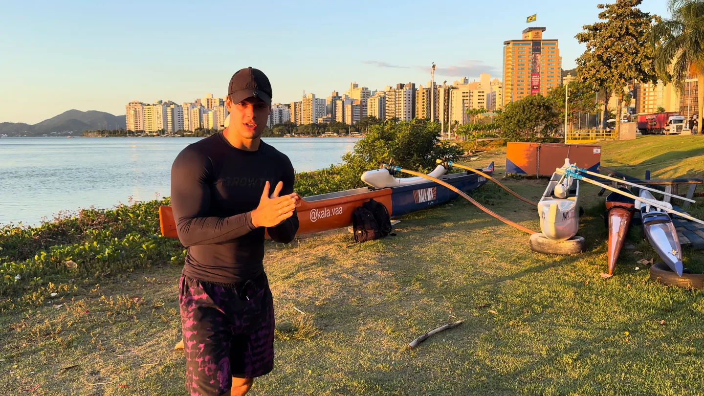 Luca Macedo, de boné e camiseta de manga comprida, falando na Beira-Mar Norte, em Florianópolis, no fim da tarde, com os prédios e a baía atrás