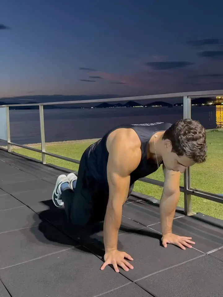 Luca Macedo, de regata, faz flexão de joelhos à beira-mar, ao anoitecer, com a baía e as luzes da cidade ao fundo.