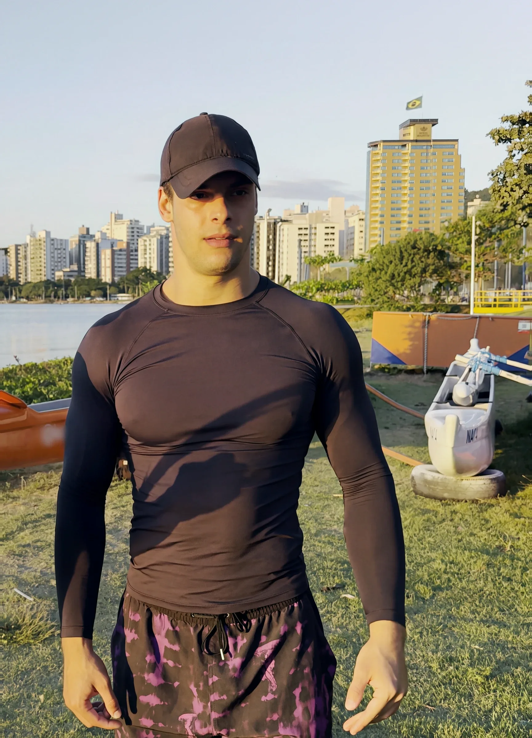 Luca Macedo, de boné e camiseta de manga comprida, em pé na Beira-Mar Norte, com os prédios e a baía atrás