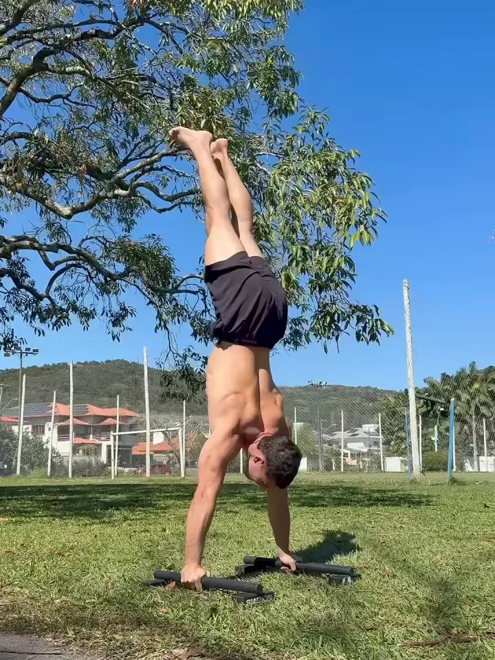 Luca Macedo em parada de mão na grama de um parque, ao sol, embaixo de uma árvore e do céu azul.